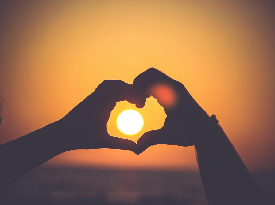 To hænder danner et hjerte foran en solnedgang.